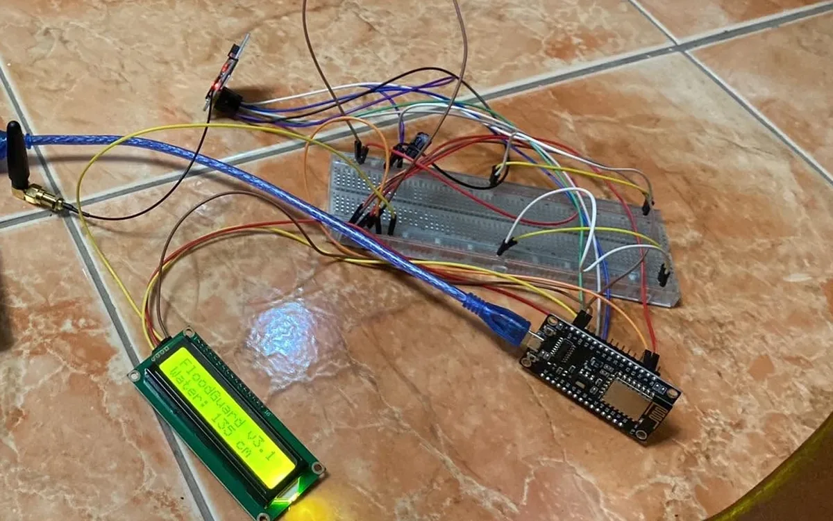 The FloodGuard prototype on a tiled floor: a NodeMCU board and a breadboard wired to a water-level sensor, a GSM module with its antenna, and a green LCD reading FloodGuard v3.1, Water: 135 cm.