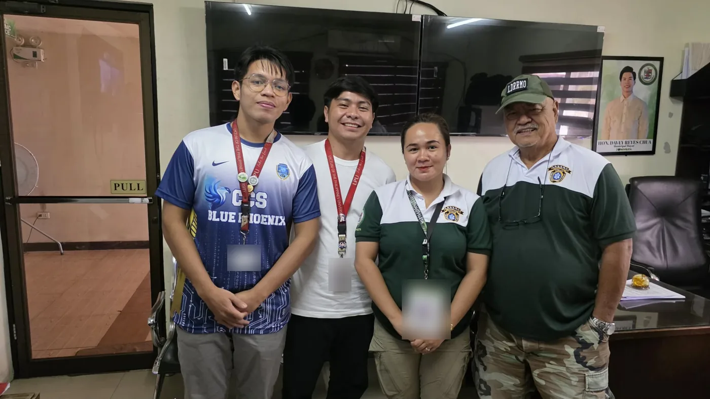 Two LPU students standing with two staff of the Noveleta local disaster risk reduction and management office in their office, a portrait of the municipal mayor on the wall behind them. The ID cards are blurred.