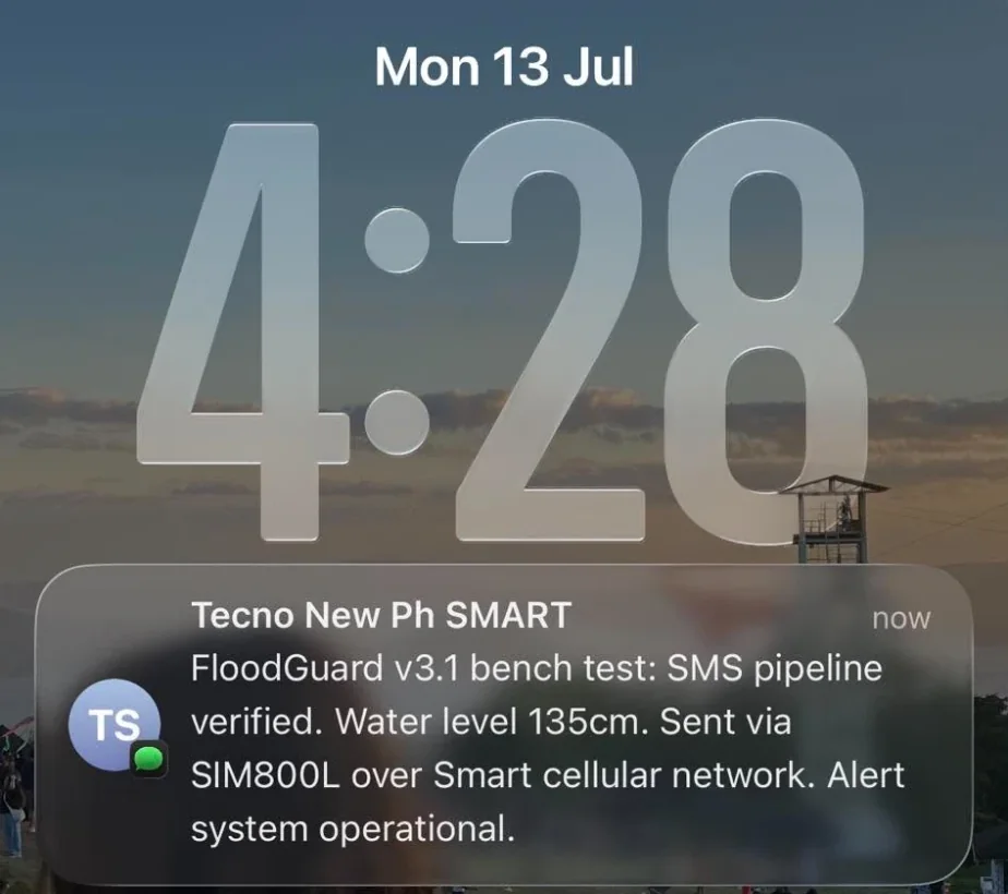 A phone lock screen on Monday 13 July at 4:28 showing a text message: FloodGuard v3.1 bench test, SMS pipeline verified, water level 135 cm, sent via SIM800L over Smart cellular network, alert system operational.