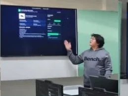A student standing beside a wall-mounted screen, one hand raised to a dark-themed web app, talking to the room.