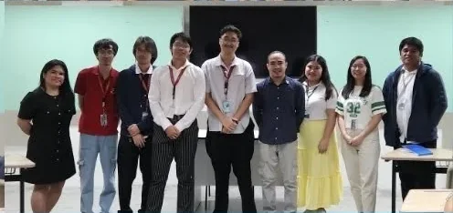Another nine people standing in a line at the front of the same classroom.